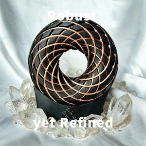 May include: A black toroidal sculpture featuring a geometric lattice pattern inlaid with copper wire. The object rests on a black base with a logo and is surrounded by clear quartz crystals. The text "Robust yet Refined" is overlaid on the image.