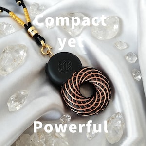 Pode incluir: Um pingente circular preto com um padrão de bobina de fio de cobre, preso a um cordão trançado preto e dourado com uma conta de metal dourado. O texto "Compact yet Powerful" está sobreposto à imagem, que apresenta pedras cristalinas.