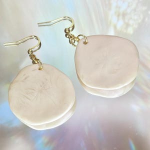 Può includere: Un paio di orecchini a strati color crema con un gancio color oro. Gli orecchini hanno una forma organica liscia con sottili dettagli superficiali. Gli orecchini sono esposti su una superficie riflettente.