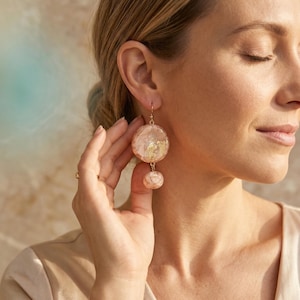May include: Gold-coloured drop earrings featuring a large, round, pinkish-white orb and a smaller, matching orb below. The orbs are filled with gold leaf flakes. The earrings are suspended from a gold hook.