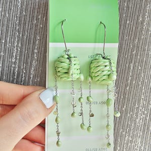 May include: Pale green earrings featuring a woven basket design and dangling beads. The earrings have long silver-coloured chains with small, oval-shaped beads in varying shades of green. The earrings are displayed against a green colour swatch.