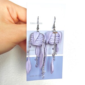 May include: A pair of lavender-coloured dangle earrings. Each earring features a woven top, dangling leaf-shaped elements, and beaded chains. The earrings are suspended from silver-coloured hooks. The text "70BB 67 096" and "82BB 38/251" are visible.
