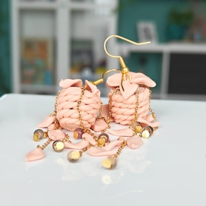 May include: Pink handmade earrings with woven basket-shaped charms, gold-coloured chain details, and dangling beads. The earrings have gold-coloured ear wires. These unique earrings are a stylish accessory, adding a touch of elegance to any outfit.