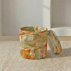 May include: A cylindrical stone container with a matching lid, featuring a polished surface with swirling patterns of green, orange, and white. The container is crafted from natural stone, displaying unique mineral veining throughout its exterior.