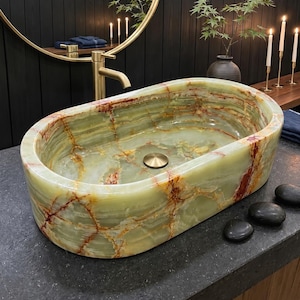 Op de afbeelding: Een ovale waskom van gepolijste groene onyx met natuurlijke aders in tinten van crème, roest en diepgroen. De waskom is voorzien van een gecentreerde ronde messing afvoerplug en gladde, afgeronde randen die de doorschijnende kwaliteit van de steen benadrukken.