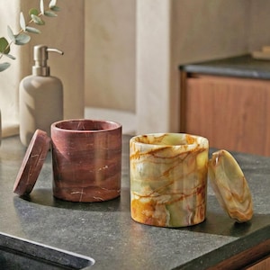 Puede incluir: Dos recipientes cilíndricos con tapas, uno rojo y otro verde, sobre una encimera oscura. Los recipientes tienen un aspecto veteado. Un dispensador de jabón y un jarrón con vegetación están en el fondo. Los recipientes son probablemente para baño o cocina.