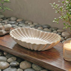 May include: A beige, oval-shaped decorative bowl with a scalloped edge sits on a wooden surface. A lit scented candle in a glass jar and a small green plant are also on the surface. The background features pebbles and a stone sculpture.
