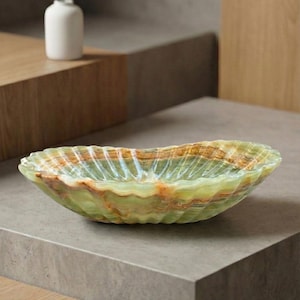 Peut inclure: Un bol décoratif de forme ovale avec un bord ondulé. Le bol est en onyx vert et marron, avec une qualité translucide. Le bol mesure environ 30 cm de long et 15 cm de large, et est posé sur une surface grise.