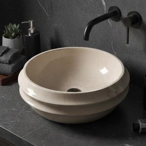 May include: A cream-colored, round bathroom sink with a tiered design. The sink is set on a dark gray countertop with a black faucet and a black soap dispenser. A small succulent, towels, and toiletries complete the modern bathroom scene.