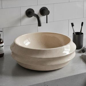 May include: A beige, round bathroom sink with a layered design. The sink is set on a gray countertop with black fixtures, including a faucet and soap dispenser. A black framed mirror hangs above the sink, with white tile in the background.