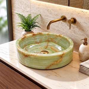 May include: A round, light green onyx stone vessel sink with natural brown veining, featuring a brass faucet and handle mounted on a beige travertine wall. A matching stone soap dispenser and a stack of folded beige towels sit on the countertop.