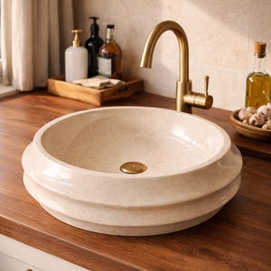 May include: A cream-colored, round bathroom sink with a decorative, tiered design. The sink has a gold-colored drain and is set on a dark wood countertop. A gold faucet is mounted above the sink. Other bathroom accessories are visible in the background.