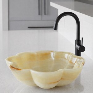 May include: A flower-shaped vessel sink carved from translucent cream and amber-toned stone, featuring smooth, rounded petals. A matte black gooseneck faucet with a single side-mounted lever handle is positioned directly behind the stone basin.