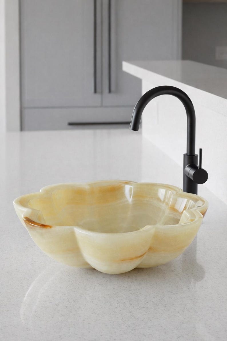 May include: A flower-shaped vessel sink carved from translucent cream and amber-toned stone, featuring smooth, rounded petals. A matte black gooseneck faucet with a single side-mounted lever handle is positioned directly behind the stone basin.