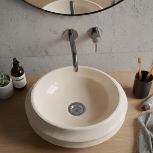 May include: Cream-colored, round bathroom sink with a chrome faucet. The sink sits on a light wood countertop, with a mirror in the background. A small succulent, soap dispenser, and toothbrush holder are also visible.