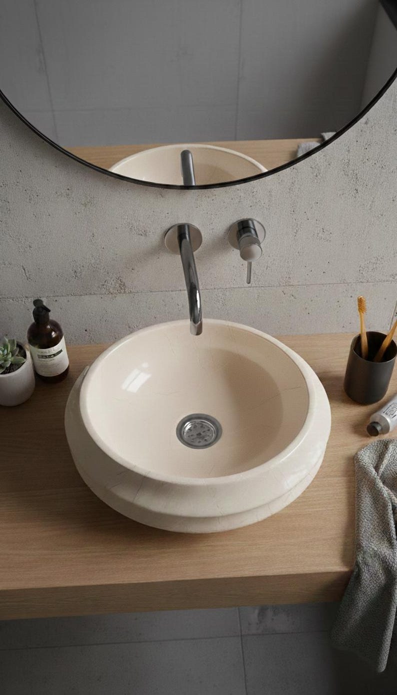 May include: Cream-colored, round bathroom sink with a chrome faucet. The sink sits on a light wood countertop, with a mirror in the background. A small succulent, soap dispenser, and toothbrush holder are also visible.