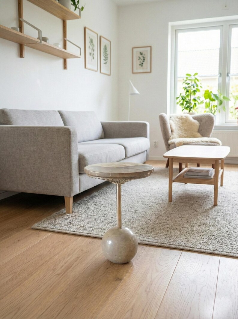 Pu&ograve; includere: Un soggiorno con un divano grigio chiaro, un tavolino rotondo con base in pietra e un tappeto colorato chiaro. La stanza ha una palette di colori neutri con pavimenti in legno e una finestra.