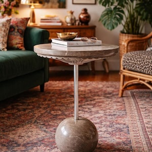 Pu&ograve; includere: Un tavolino rotondo con piano in marmo grigio e base sferica. Il tavolo ha un gambo in metallo argentato. Sopra il tavolo ci sono libri, una ciotola dorata e occhiali. Il tavolo &egrave; in una stanza con un tappeto fantasia.
