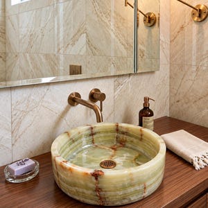 May include: A round vessel sink crafted from light green onyx stone with natural brown veining. The sink features a polished finish and a circular bronze drain cover, paired with a wall-mounted bronze faucet fixture above the basin.