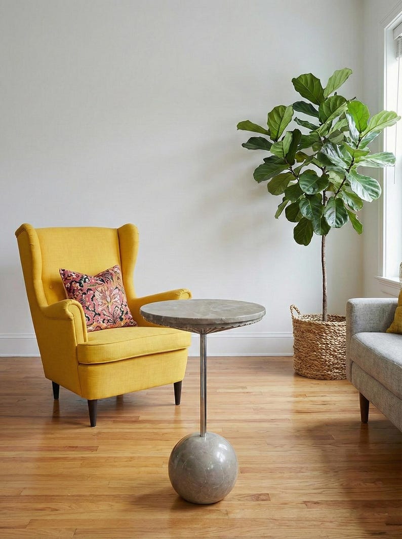 May include: A yellow armchair with a patterned pillow sits next to a small, round side table with a gray top and a spherical base. A large potted plant with green leaves is in a woven basket, adding a touch of nature to the room.