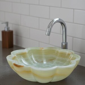 May include: A flower-shaped vessel sink carved from pale green and translucent white onyx stone with natural golden-brown veining. The basin features a scalloped rim and a smooth, polished finish, paired with a curved chrome faucet.