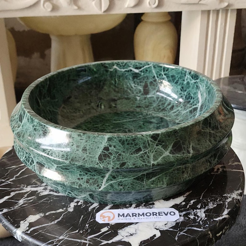 May include: A round, green marble bowl with a decorative, ridged exterior. The bowl sits on a black and white marble surface. The bowl's interior is smooth and polished, showcasing the natural veining of the stone. The logo "MARMOREVO Marble & Handicrafts" is visible.
