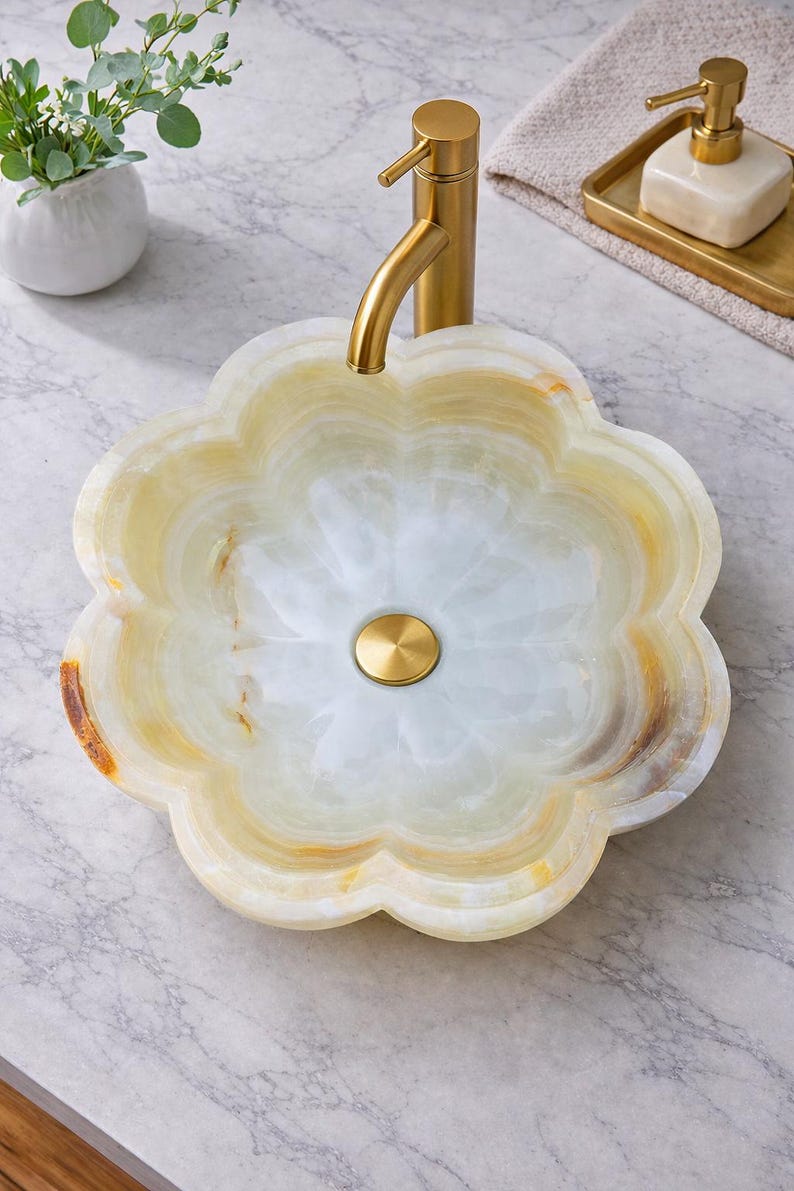 May include: A flower-shaped bathroom sink made of natural stone with a gold-colored faucet. The sink has a light green and beige color scheme with a gold drain. The sink is a unique and stylish addition to any bathroom.