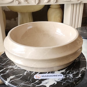 May include: A beige marble basin with a rounded shape and a layered, textured exterior. The basin sits on a black and white marble surface. The logo "MARMOREVO Marble & Handicrafts" is visible.