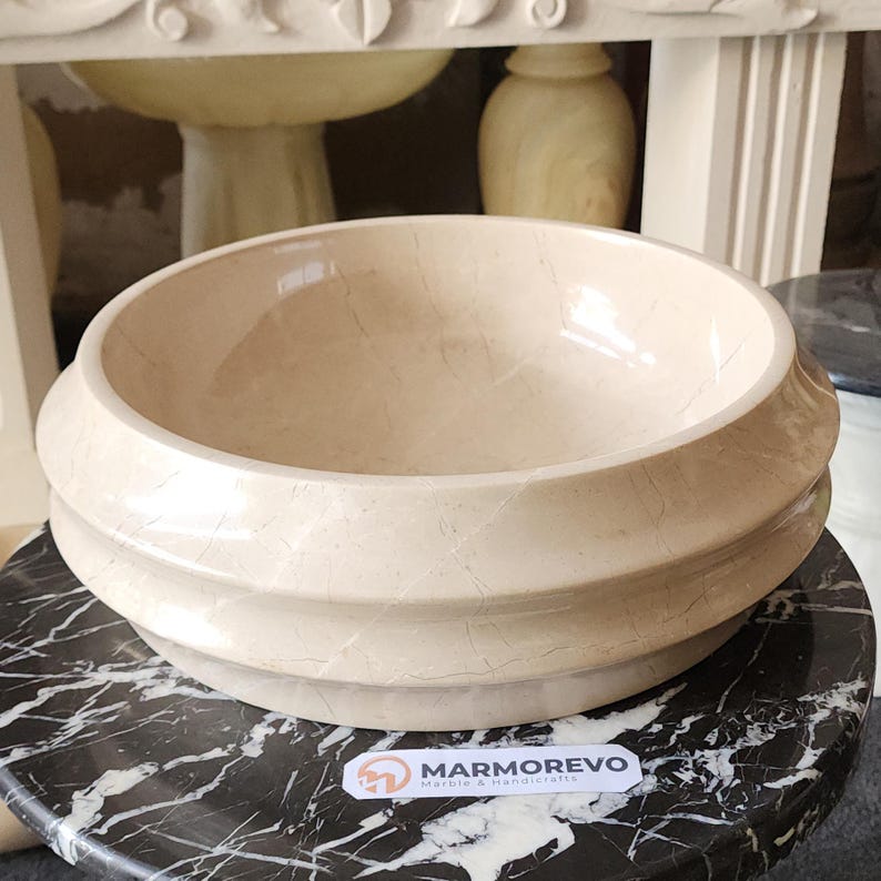 May include: A beige marble basin with a rounded shape and a layered, textured exterior. The basin sits on a black and white marble surface. The logo "MARMOREVO Marble & Handicrafts" is visible.