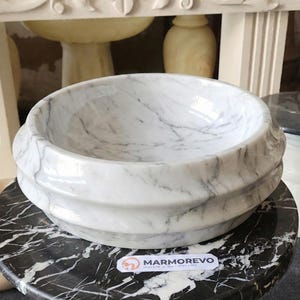 May include: A white marble bowl with gray veining, featuring a layered, ridged design. The bowl sits on a black and white marble surface. The logo "MARMOREVO Marble & Handicrafts" is visible.