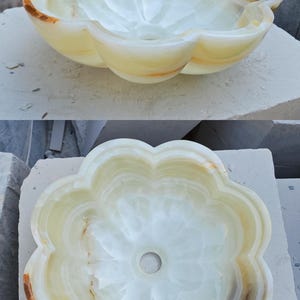May include: A flower-shaped vessel sink carved from light green and cream-colored onyx stone. The basin features smooth, rounded petals and a central drain hole, showcasing natural translucent veining and warm amber-toned mineral inclusions.
