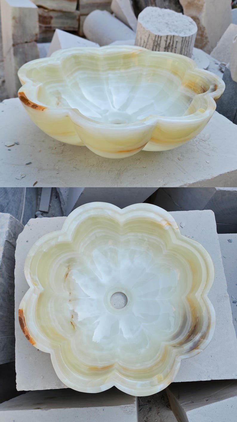 May include: A flower-shaped vessel sink carved from light green and cream-colored onyx stone. The basin features smooth, rounded petals and a central drain hole, showcasing natural translucent veining and warm amber-toned mineral inclusions.