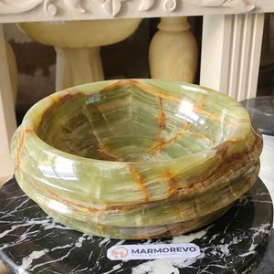 May include: A green onyx stone basin with brown veining, displayed on a black and white marble table. The basin has a rounded shape with a polished surface. The background includes decorative elements.