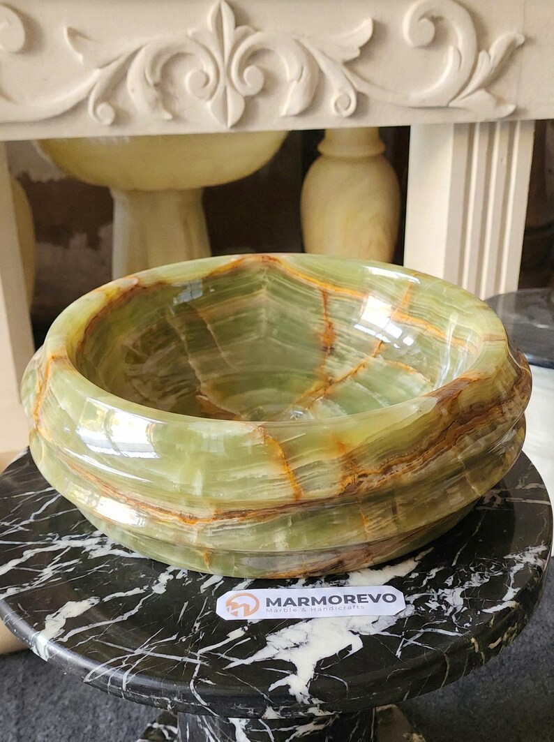 May include: A green onyx stone basin with brown veining, displayed on a black and white marble table. The basin has a rounded shape with a polished surface. The background includes decorative elements.