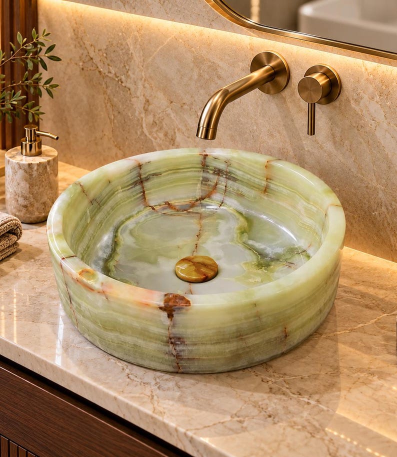 May include: A round vessel sink crafted from polished green onyx stone with natural brown veining. The sink features a matching stone drain cover and is paired with a wall-mounted brushed gold faucet and a single lever handle control.