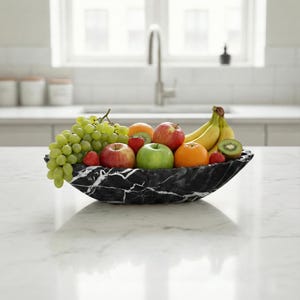 May include: A black marble fruit bowl filled with a variety of fresh fruit, including green grapes, apples, oranges, strawberries, bananas, and kiwi. The bowl has a unique shape and sits on a white countertop in a kitchen setting.