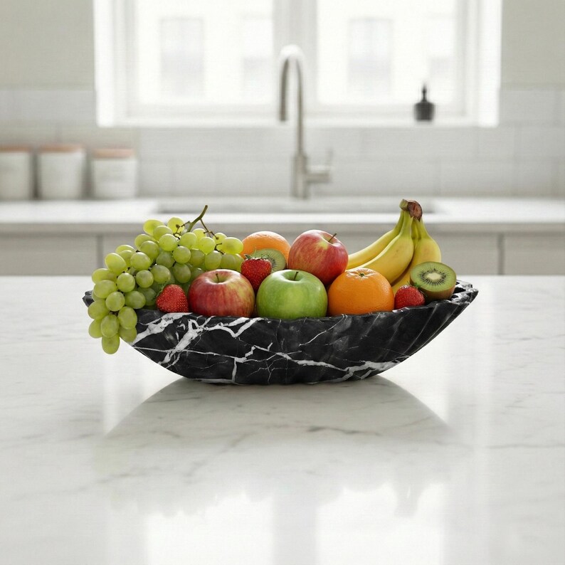 May include: A black marble fruit bowl filled with a variety of fresh fruit, including green grapes, apples, oranges, strawberries, bananas, and kiwi. The bowl has a unique shape and sits on a white countertop in a kitchen setting.