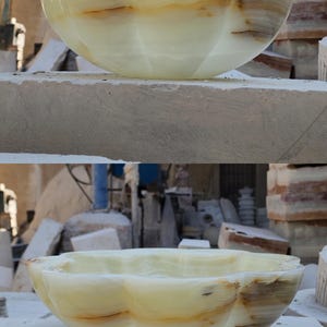May include: A scalloped bowl carved from pale green onyx stone featuring natural translucent layers and brown mineral veining. The vessel has a smooth, polished finish with a curved, flower-like rim and a deep, rounded basin structure.