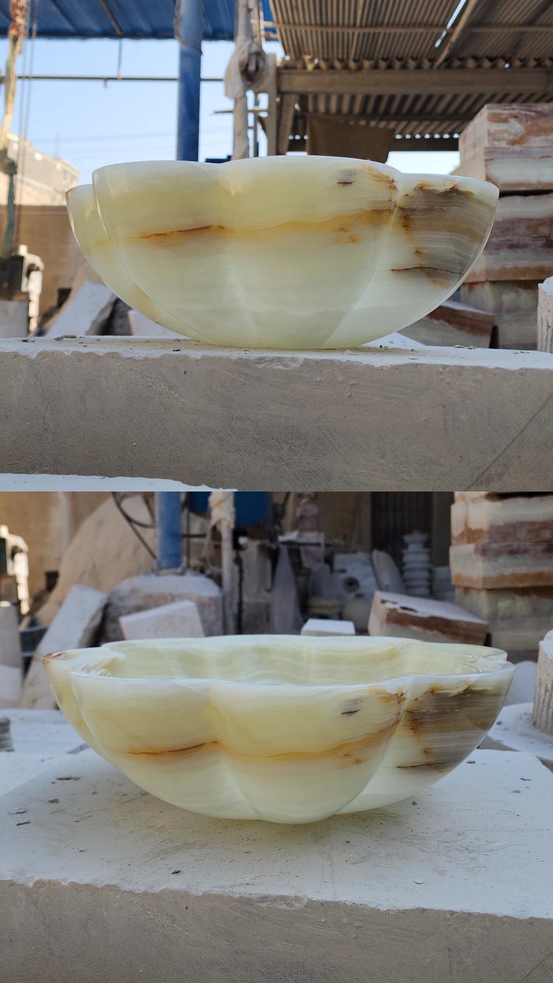 May include: A scalloped bowl carved from pale green onyx stone featuring natural translucent layers and brown mineral veining. The vessel has a smooth, polished finish with a curved, flower-like rim and a deep, rounded basin structure.