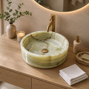May include: A round, light green onyx stone vessel sink with natural veining and a polished finish. The sink features a central drain and is paired with a tall, brushed brass faucet. The basin sits on a wooden vanity surface.