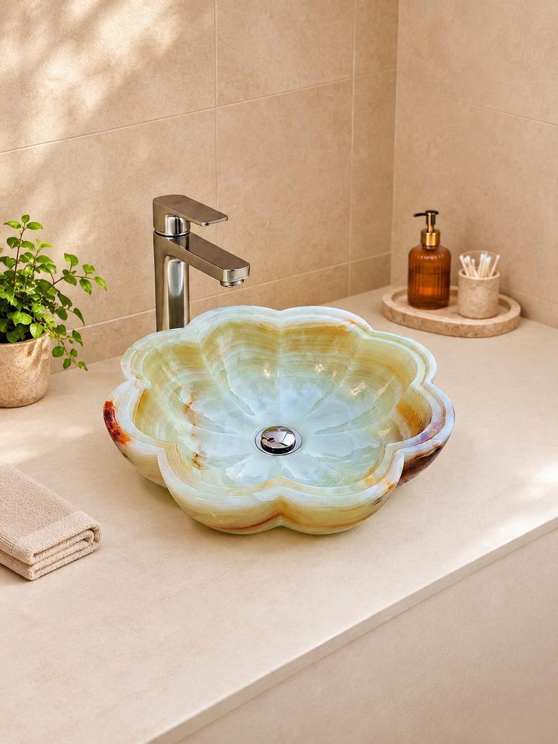 May include: A flower-shaped vessel sink carved from translucent green and amber onyx stone, featuring a polished chrome drain stopper and a tall, brushed nickel faucet mounted on a smooth, light-colored countertop surface.