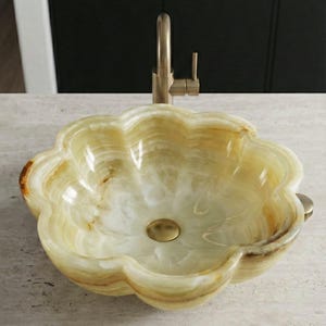 May include: A flower-shaped onyx sink with a brass faucet. The sink has a light green and cream color with brown veining. The faucet and drain are brass-colored. The sink is on a light-colored countertop.