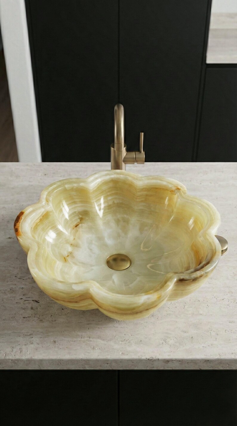 May include: A flower-shaped onyx sink with a brass faucet. The sink has a light green and cream color with brown veining. The faucet and drain are brass-colored. The sink is on a light-colored countertop.
