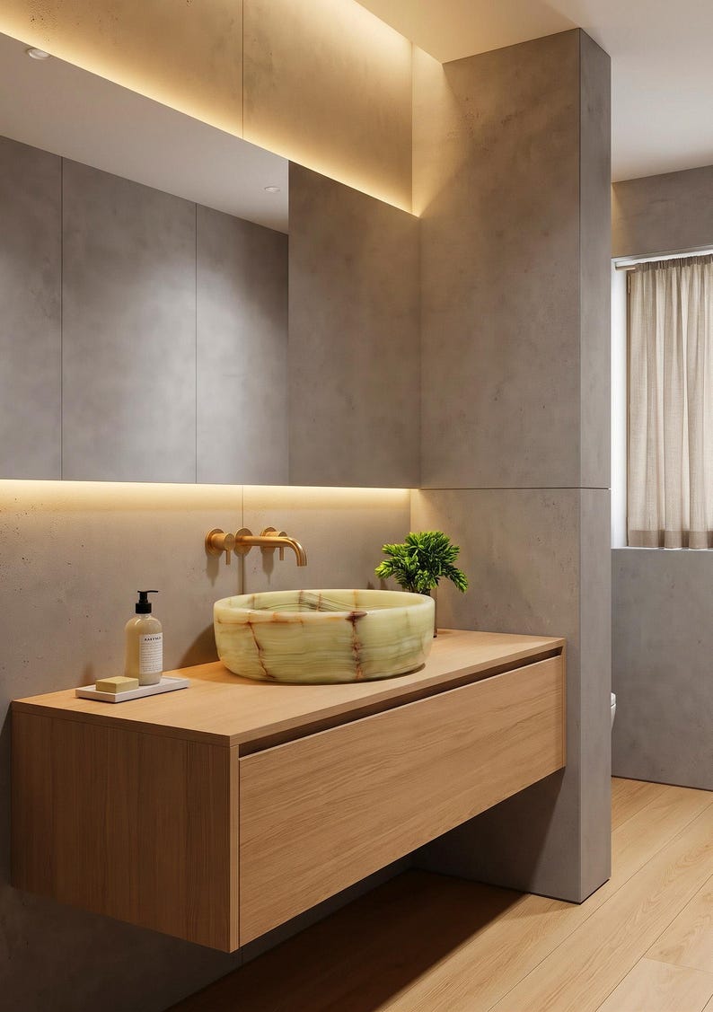 May include: A round, light green onyx vessel sink with natural brown veining sits atop a light wood floating vanity. A wall-mounted brass tap is positioned above the sink, accompanied by a soap dispenser and a small green plant.