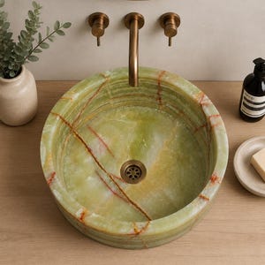 May include: A round vessel sink crafted from polished green onyx stone featuring natural orange and brown veining throughout. The basin includes a central bronze-toned drain and is paired with a matching wall-mounted bronze faucet fixture.