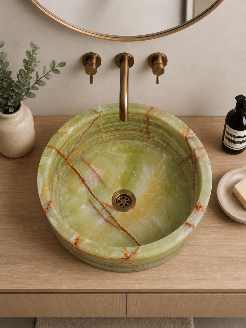 May include: A round vessel sink crafted from polished green onyx stone featuring natural orange and brown veining throughout. The basin includes a central bronze-toned drain and is paired with a matching wall-mounted bronze faucet fixture.