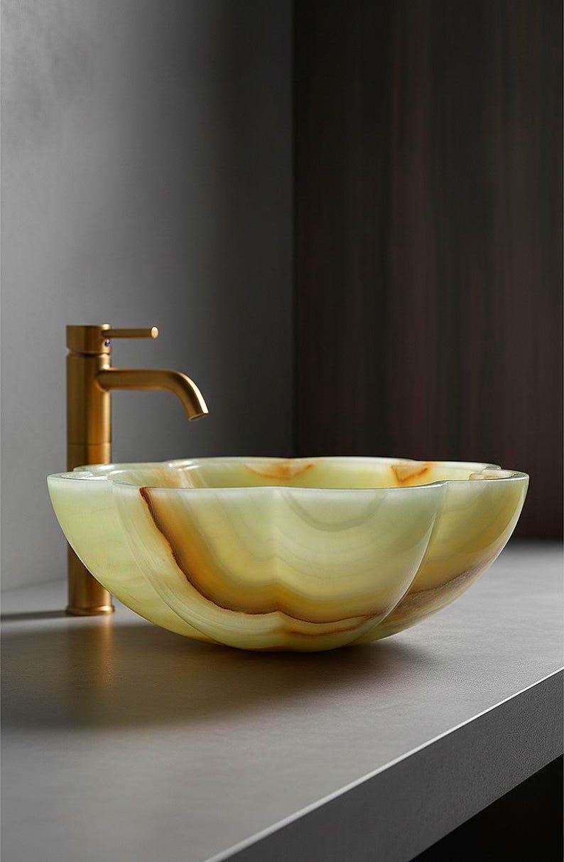 May include: A scalloped vessel sink carved from translucent light green and amber onyx stone, featuring natural banded patterns throughout the material. A tall, brushed gold-toned metal faucet stands directly behind the basin.