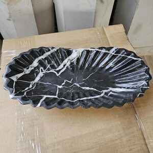 May include: A black marble oval serving dish with white veining. The dish has a scalloped edge and a textured, fan-like pattern on the interior. The dish is sitting on a cardboard surface.