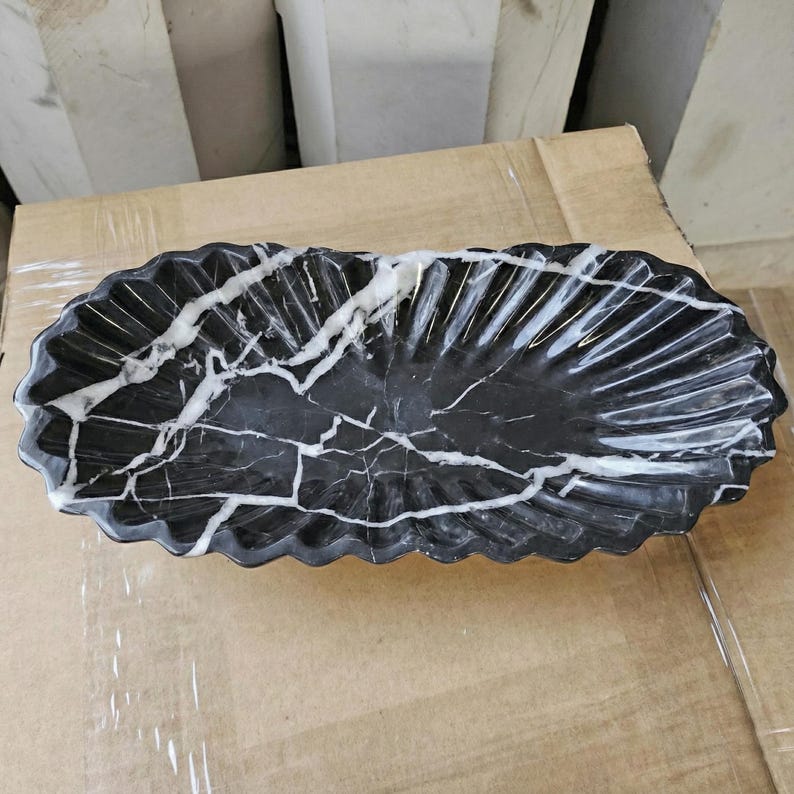 May include: A black marble oval serving dish with white veining. The dish has a scalloped edge and a textured, fan-like pattern on the interior. The dish is sitting on a cardboard surface.