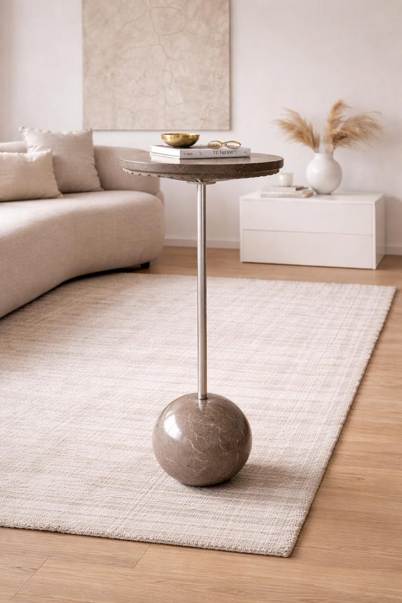 Pu&ograve; includere: Un tavolino moderno con un piano rotondo e strutturato e uno stelo in metallo lucido. La base &egrave; di forma sferica con un aspetto marmorizzato. Il tavolo &egrave; appoggiato su un tappeto colorato chiaro in una stanza dai toni neutri.